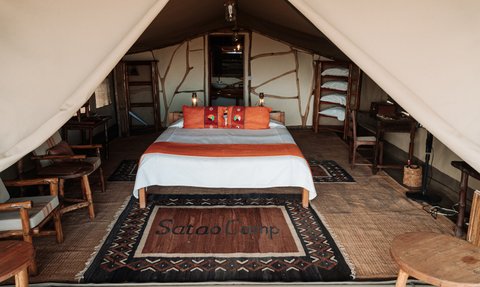 Ein gemütliches Zeltinnere mit einem großen Bett, bunten Kissen und einem Teppich mit der Aufschrift 'Satao Camp'.
