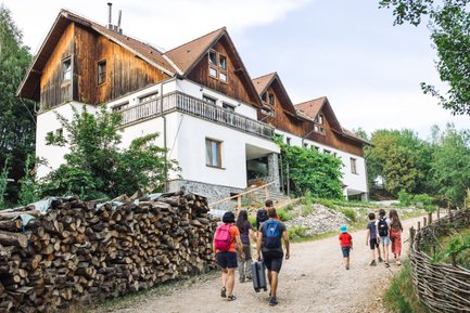 Eine Gruppe von fünf Personen geht auf einem Weg zu einem großen, rustikalen Haus, umgeben von Bäumen und Holzstapeln.