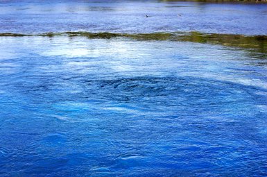 Ein ruhiger Wasserbereich mit sanften Wellen und einem Wirbel in der Mitte, umgeben von blauen und grünen Farbtönen.