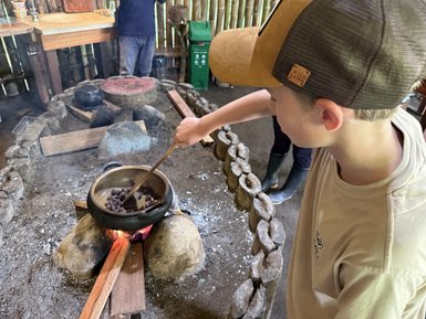 Ein Junge rührt mit einem Holzlöffel in einem Topf über einem offenen Feuer, während er konzentriert zusieht.