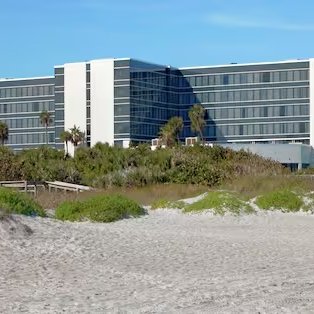 Ein modernes Hotelgebäude steht direkt am Strand, umgeben von üppigem Grün und Palmen, unter klarem blauen Himmel.