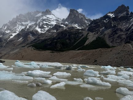 Im Vordergrund schwimmen Eisblöcke in einem trüben Gewässer, während majestätische Berge im Hintergrund aufragen.