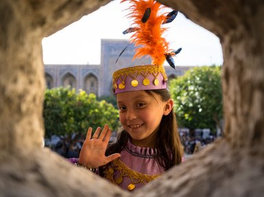 Ein fröhliches Mädchen mit einem bunten Hut und einer Feder winkt durch ein Fenster, umgeben von grünen Bäumen.