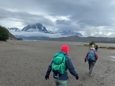 Eine Gruppe von Wanderern geht über einen grauen Sandstrand, umgeben von majestätischen Bergen und bewölktem Himmel.