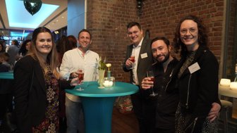 Eine Gruppe von fünf Personen lächelt an einem Tisch mit Getränken, umgeben von einer festlichen Atmosphäre.