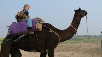 Ein fröhlicher Junge sitzt auf einem Kamel, während er mit einer Hand winkt und in die Kamera lächelt.