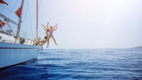 Drei Kinder springen fröhlich von einer Yacht ins glitzernde blaue Wasser, umgeben von einem klaren Himmel.