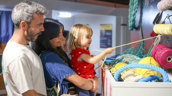 Eine Familie betrachtet farbige Seile und Netze in einem Ausstellungsraum, während ein kleines Mädchen mit einem Seil spielt.