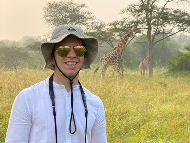 Ein junger Mann mit Sonnenbrille und Hut steht in einer grünen Savanne, während Giraffen im Hintergrund grasen.