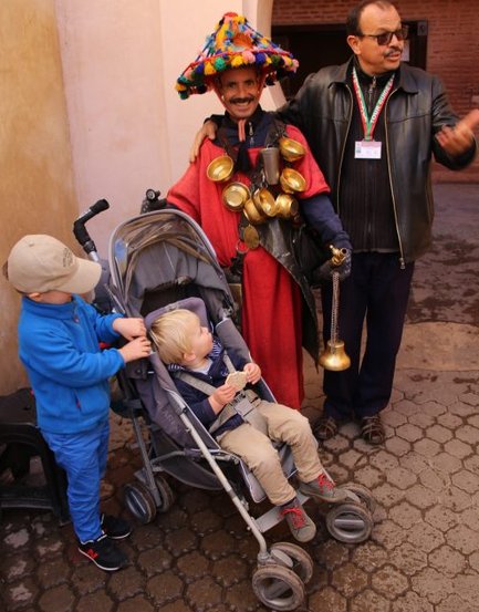 Ein fröhlicher Mann in traditioneller Kleidung mit einem bunten Hut steht neben zwei Kindern in einem gepflasterten Bereich.