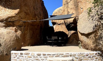 Eine gemütliche Sitzecke unter einem Sonnensegel, umgeben von beeindruckenden Felsen und klarem blauen Himmel.