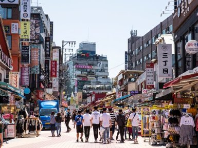 Eine belebte Einkaufsstraße in Hongdae, gesäumt von Geschäften und bunten Schildern, mit vielen Passanten im Sonnenlicht.