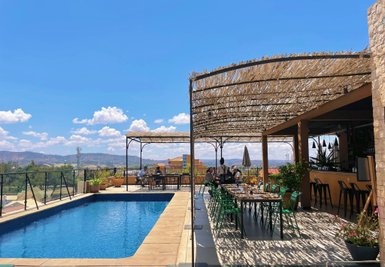 Ein sonniger Poolbereich mit einem Restaurant im Freien, umgeben von Pflanzen und Blick auf die Berge im Hintergrund.