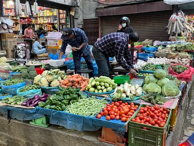 Zwei Verkäufer sortieren frisches Gemüse auf einem belebten Markt in Thamel, umgeben von bunten Ständen und Gebäuden.