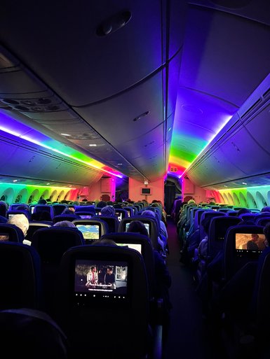 Der Innenraum eines Flugzeugs ist in bunten Lichtern beleuchtet, während Passagiere Filme auf Bildschirmen ansehen.