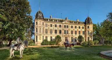 Ein prächtiges historisches Gebäude mit Türmen und einem gepflegten Garten, in dem zwei Reiter auf ihren Pferden stehen.