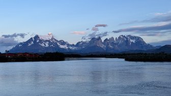 Die majestätischen Torres del Paine erheben sich über einen ruhigen Fluss, während die Wolken sanft am Himmel ziehen.