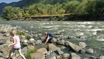 Ein Mädchen mit langen Haaren läuft barfuß über große Steine am Ufer eines rauschenden Flusses in einer grünen Landschaft.