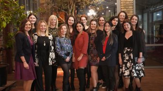 Eine Gruppe von Frauen posiert fröhlich in einem stilvollen Raum mit Pflanzen und warmem Licht im Hintergrund.