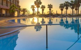 Ein atemberaubender Sonnenuntergang spiegelt sich im glitzernden Pool eines Hotels, umgeben von Palmen und Liegen.