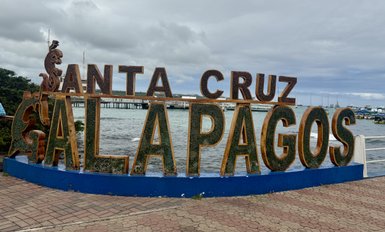 Der bunte Schriftzug 'Santa Cruz Galapagos' steht am Ufer, umgeben von Wasser und einem bewölkten Himmel.