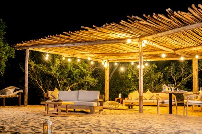 Ein gemütlicher Strandbereich mit einem Holzunterstand, beleuchtet von warmen Lichtern, umgeben von grünen Bäumen.