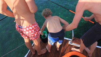 Ein Junge in Badehose beugt sich über die Reling eines Bootes und greift nach einem Eimer im klaren Wasser.