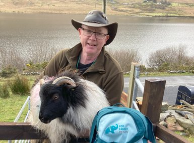 Ein lächelnder Mann mit Brille trägt einen Hut und hält ein Schaf auf einem Zaun, während eine malerische Landschaft im Hintergrund zu sehen ist.
