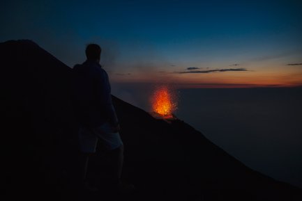 Ein Mensch steht am Rand eines Vulkans und beobachtet die beeindruckende Eruption mit glühender Lava und Funken im Abendlicht.