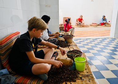Kind hilft einheimischen Frauen beim Mahlen von Arganfrüchten zur Ölgewinnung in Essaouira – Marokko Familienreise