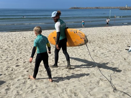 Ein Surflehrer und ein Junge gehen am Strand entlang, während der Lehrer ein großes, orangefarbenes Surfbrett trägt.