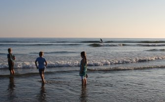 Drei Kinder stehen im flachen Wasser, während sanfte Wellen an den Strand rollen und die Sonne am Horizont scheint.