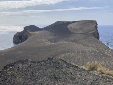 Eine beeindruckende Küstenlandschaft mit schroffen Vulkanhängen und glitzerndem Wasser im Hintergrund, unter einem bewölkten Himmel.