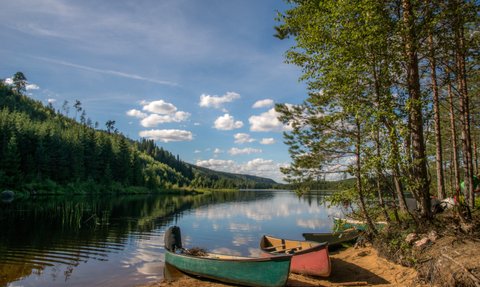 Ein ruhiger See umgeben von dichten Wäldern, mit bunten Kanus am Ufer, die auf das glitzernde Wasser blicken.
