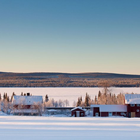 Eine verschneite Winterlandschaft mit roten Holzhäusern, umgeben von schneebedeckten Bäumen und sanften Hügeln im Hintergrund.