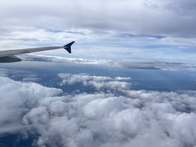 Der Blick aus dem Flugzeug zeigt die Wolkenlandschaft unterhalb, mit einem Flugzeugflügel im Vordergrund.