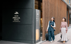 Zwei Frauen stehen vor dem Keio Prelia Hotel, lächelnd und mit Gepäck, bereit für ihren Aufenthalt in Kyoto.