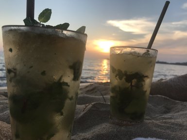 Zwei erfrischende Cocktails mit Minze stehen auf dem Sandstrand, während die Sonne am Horizont untergeht.