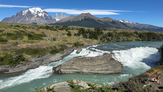 Ein beeindruckender Wasserfall stürzt über schichtige Felsen, umgeben von üppiger Vegetation und majestätischen Bergen im Hintergrund.