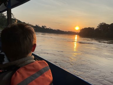 Ein Junge in einem orangefarbenen Schwimmwesten blickt auf den Sonnenuntergang über einem ruhigen Fluss in der Amazonasregion.