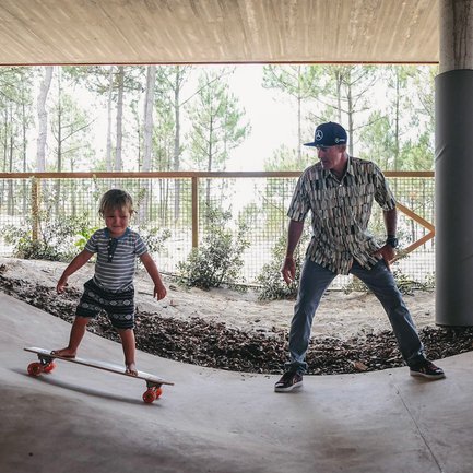 Ein kleiner Junge steht auf einem Skateboard und lächelt, während ein Erwachsener ihn ermutigend beobachtet.