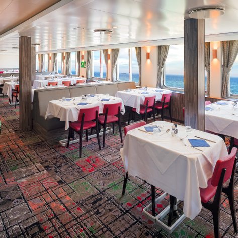 Ein eleganter Essbereich auf einem Schiff mit weißen Tischdecken und Blick auf das offene Meer durch große Fenster.