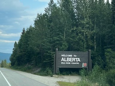 Ein Willkommensschild für Alberta, umgeben von üppigem Grün und hohen Bäumen, unter einem bewölkten Himmel.