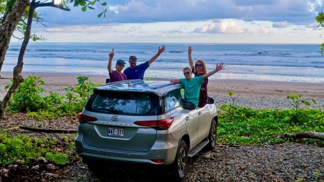 Eine Gruppe von vier Personen steht fröhlich neben einem silbernen Auto am Strand von Costa Rica, umgeben von üppigem Grün.