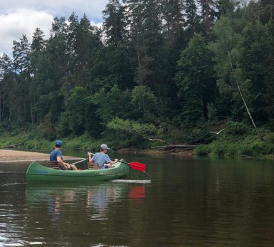 Zwei Personen paddeln in einem grünen Kanu auf einem ruhigen Fluss, umgeben von üppigem Wald und bewölktem Himmel.