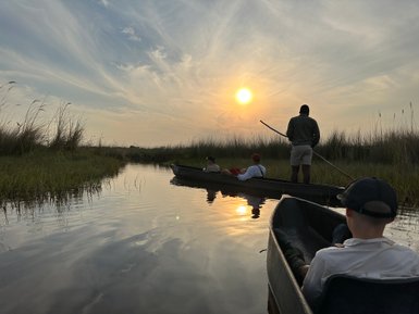 Eine Gruppe von Menschen paddelt in Kanus durch eine ruhige Wasserstraße, umgeben von hohem Gras und einem malerischen Sonnenuntergang.