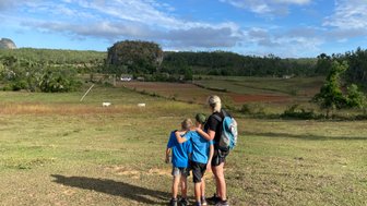 Eine Frau steht mit zwei Kindern auf einer Wiese und blickt auf eine malerische Landschaft mit sanften Hügeln.