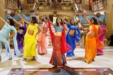 Eine Gruppe von Tänzerinnen in bunten traditionellen Outfits führt einen lebhaften Bollywood-Tanz in einem festlichen Saal auf.