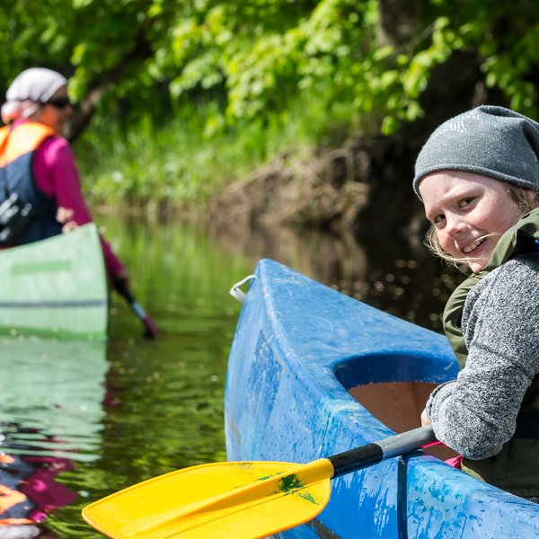 Ein fröhliches Kind paddelt in einem blauen Kajak auf einem ruhigen Gewässer, umgeben von grüner Natur und anderen Kajakfahrern.