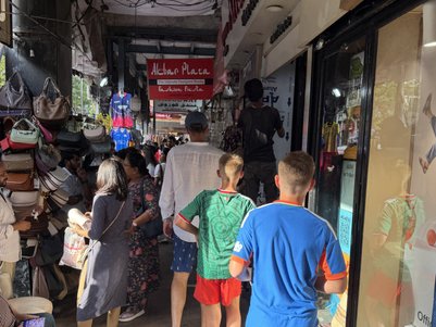 Zwei Jungen in bunten T-Shirts gehen durch einen belebten Markt in Mumbai, umgeben von Geschäften und Menschen.